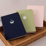 Tsuki ‘Cup of Galaxy’ Limited Edition Holographic Bullet Journal with Tsuki ‘Ichigo’ Limited Edition Boba Bullet Journal and Tsuki ‘Matcha Matcha’ Limited Edition notebook in brown basket on beige linen covered table 