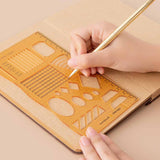 Tsuki Bullet Journal Stencil Set in neutral demonstrated on open bullet journal kraft paper page spread with pencil held in hands on beige background