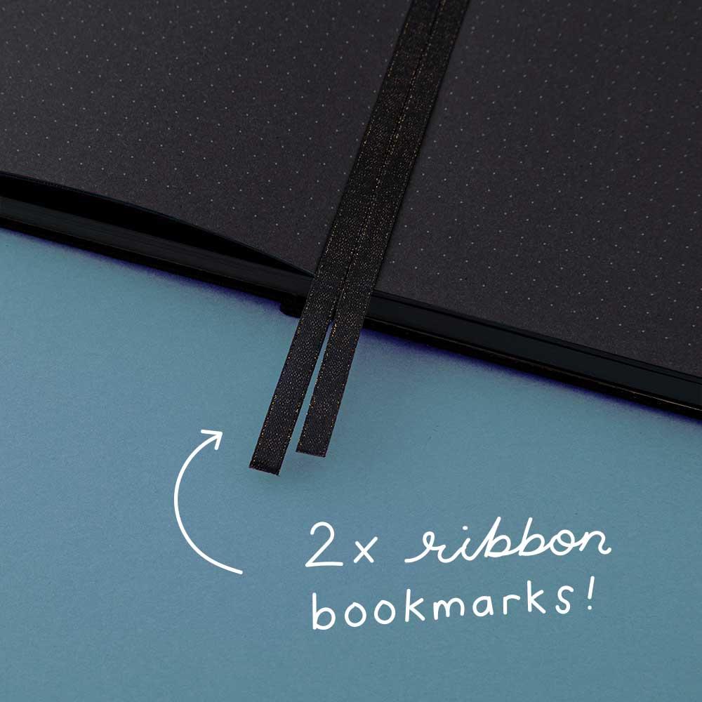 Tsuki Black Paper Limited Edition Hardcover Bullet Journal in Moonflower held in hands at spine angle in dark blue background