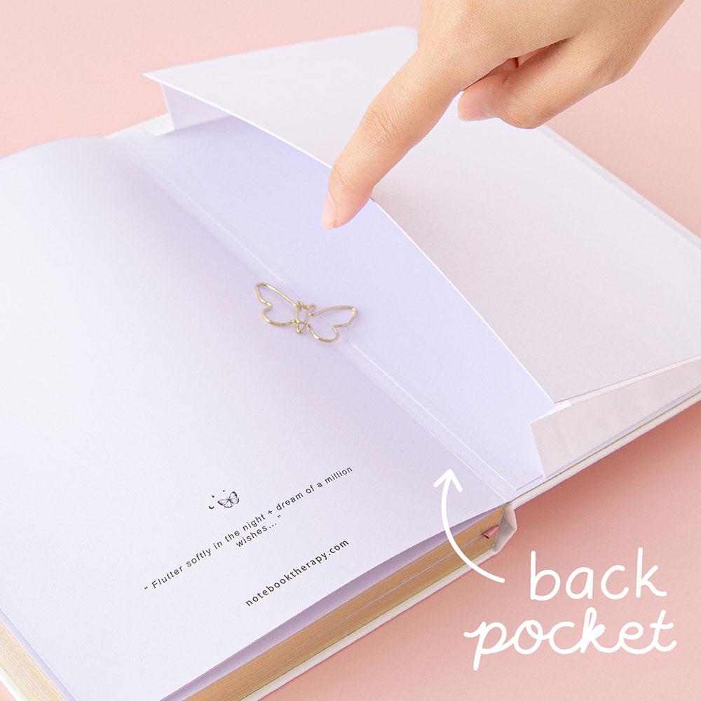 Close up of open back page pocket of Tsuki Cloud White ‘Flutter + Dream’ Limited Edition Bullet Journal by Notebook Therapy x Pelinkan with free butterfly bookmark gift held in hand on pastel pink background